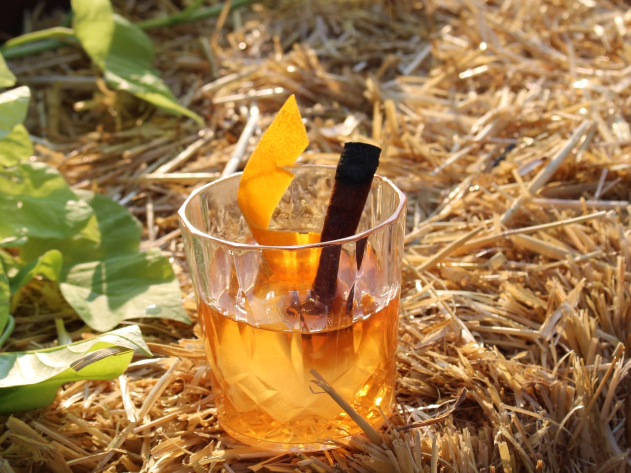 Golden bourbon cocktail with an orange peel and charred cinnamon stick, served on hay bales in the evening sun.