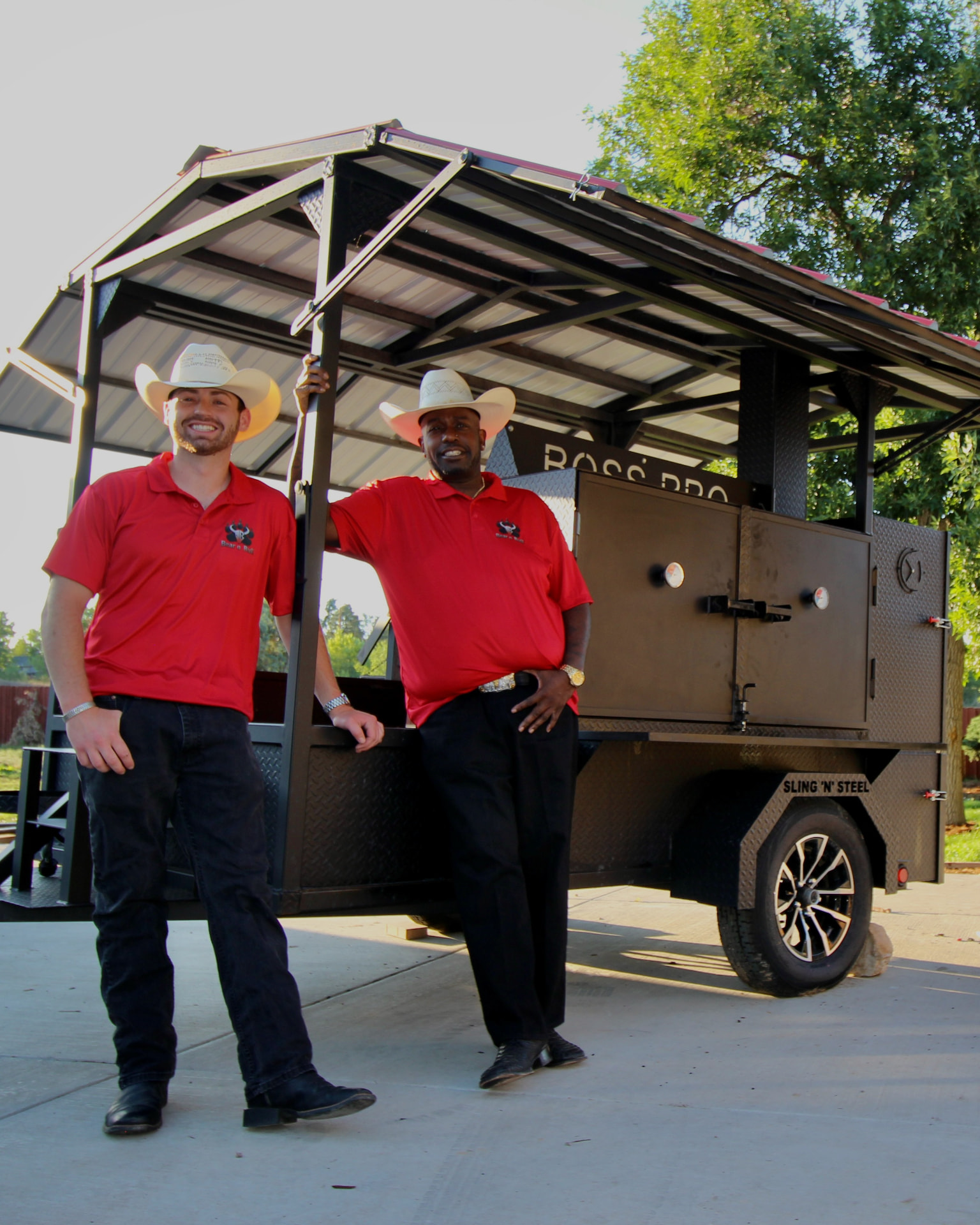 Will and Ryan in Bear n Bull red shirts and cowboy hats standing beside the smoker trailer.